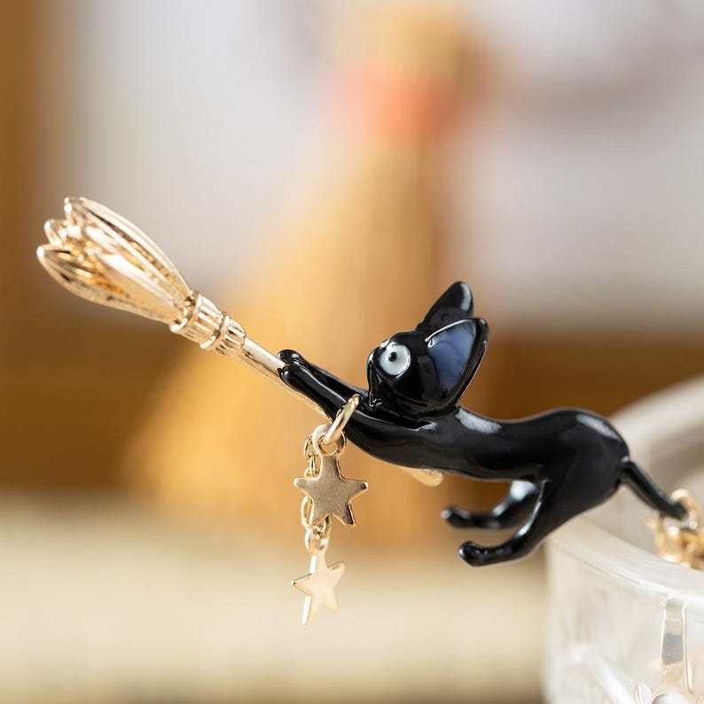May include: A black cat brooch with a gold-colored broom and dangling star charms. The cat is in a flying pose, with its eyes wide open. The brooch is likely made of enamel and metal.
