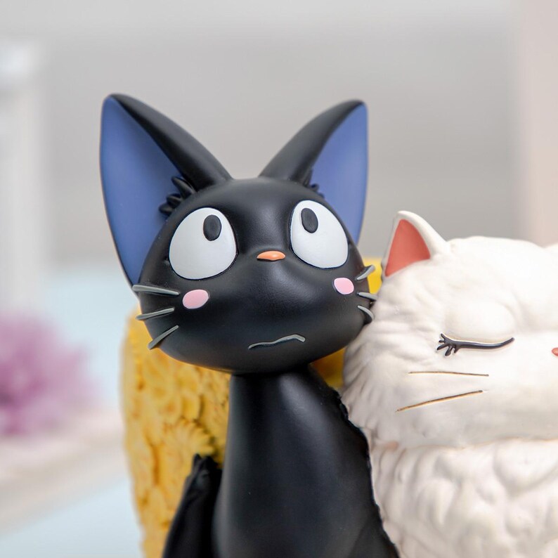 Puede incluir: Una figura que muestra un gato negro con grandes ojos blancos, orejas interiores azules y manchas rosas en las mejillas, junto a un gato blanco con los ojos cerrados y pelaje texturizado. El gato negro tiene una peque&ntilde;a nariz naranja.