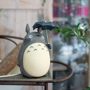 Puede incluir: Una figura de Totoro de color gris y crema con cuerpo redondeado, orejas puntiagudas y bigotes. El personaje sostiene un peque&ntilde;o paraguas negro abierto con un mango plateado. La figura tiene un acabado mate liso y una barriga clara.