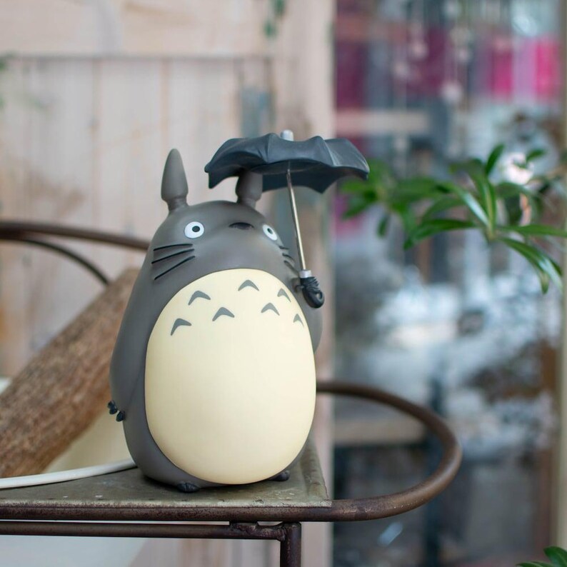 Puede incluir: Una figura de Totoro de color gris y crema con cuerpo redondeado, orejas puntiagudas y bigotes. El personaje sostiene un peque&ntilde;o paraguas negro abierto con un mango plateado. La figura tiene un acabado mate liso y una barriga clara.