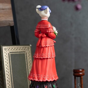 Puede incluir: Figura decorativa de una dama gato con un vestido rojo, sosteniendo flores blancas. La figura lleva un sombrero azul y un collar de perlas. El vestido tiene un dise&ntilde;o escalonado con detalles blancos. La figura est&aacute; sobre una base decorativa.