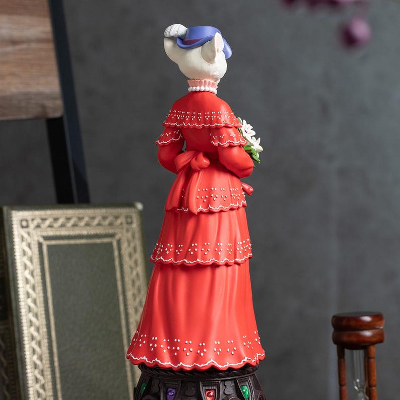 Puede incluir: Figura decorativa de una dama gato con un vestido rojo, sosteniendo flores blancas. La figura lleva un sombrero azul y un collar de perlas. El vestido tiene un dise&ntilde;o escalonado con detalles blancos. La figura est&aacute; sobre una base decorativa.