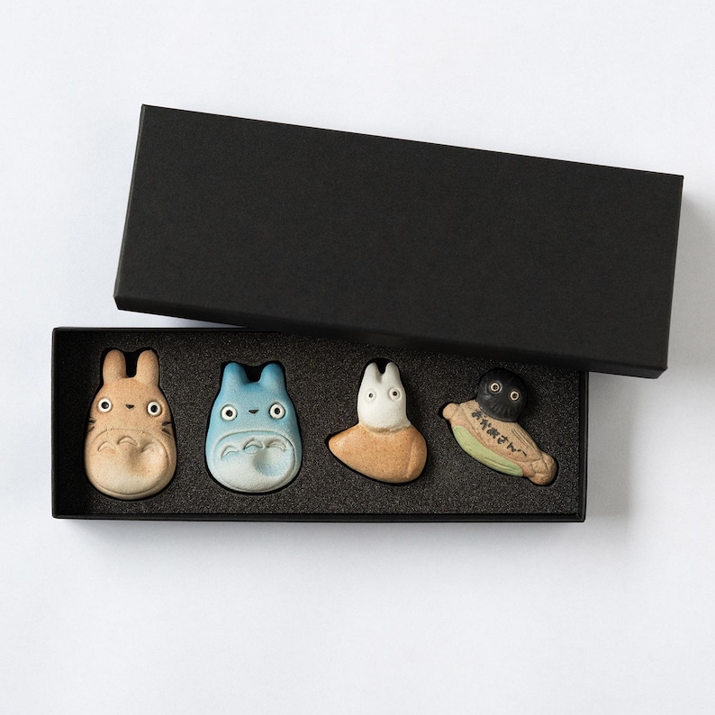 Puede incluir: Un conjunto de cuatro peque&ntilde;as figuras de cer&aacute;mica dispuestas en una caja de regalo rectangular negra con tapa a juego. Las figuras incluyen una criatura beige, una azul, una blanca y una negra y verde con texto japon&eacute;s en el cuerpo.