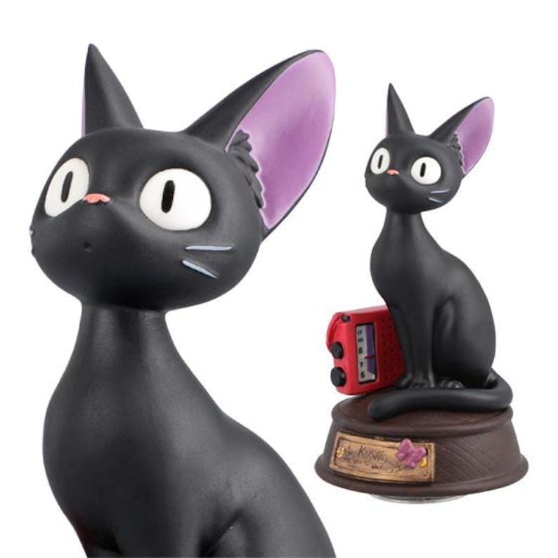 May include: A black cat figurine with large white eyes, pink nose, and purple-lined ears. The cat is sitting on a brown base with a red device. The figurine has a cartoonish design.