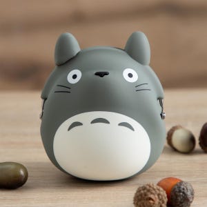 May include: A gray Totoro-shaped coin purse with a white belly and black facial details. The purse has two small ears and a zipper closure. Acorns are scattered around the purse on a wooden surface.