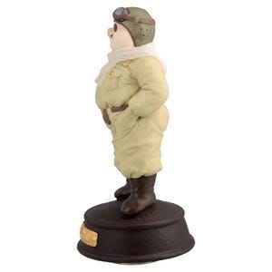 May include: A ceramic figurine of a pig in a vintage aviator outfit. The pig is wearing a khaki flight suit, brown boots, and a green aviator helmet with goggles. A white scarf is around its neck. The figurine stands on a dark brown base.