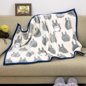 Puede incluir: Una manta de felpa color crema con un estampado de personajes Totoro grises. La manta tiene un borde azul oscuro y está colocada sobre un sofá. Una imagen enmarcada y ovillos de lana son visibles en el fondo.