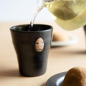 Könnte beinhalten: Eine schwarze Keramiktasse mit einem geschnitzten Holzgesicht-Design wird mit einer klaren Flüssigkeit gefüllt. Die Tasse steht auf einer hellen Holzoberfläche. Die Tasse hat eine zylindrische Form und eine glatte, matte Oberfläche.