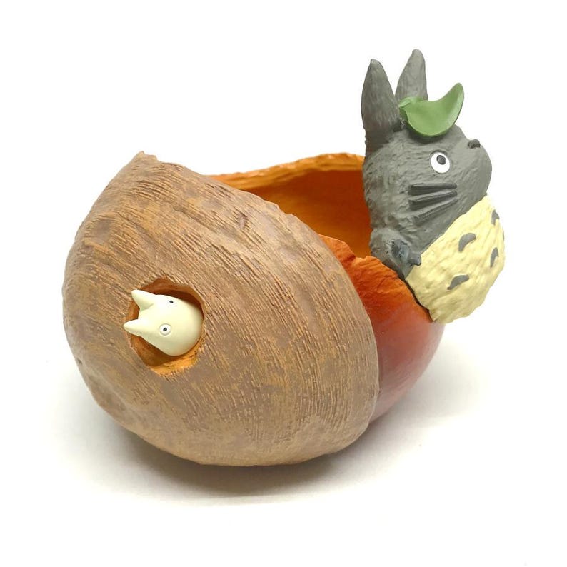 K&ouml;nnte beinhalten: Ein dekorativer Pflanztopf mit einer grauen Totoro-Figur mit gr&uuml;nem Blatthut, die aus einem braunen, ausgeh&ouml;hlten Baumstamm herausschaut. Eine kleine wei&szlig;e Kreatur ist in einem Loch an der Seite sichtbar. Der Topf hat einen orangefarbenen Innenraum.