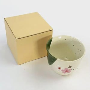 May include: A ceramic teacup with a white and green design, featuring pink floral accents and black speckles. The teacup is next to a beige cardboard box. The teacup has a unique, asymmetrical shape.