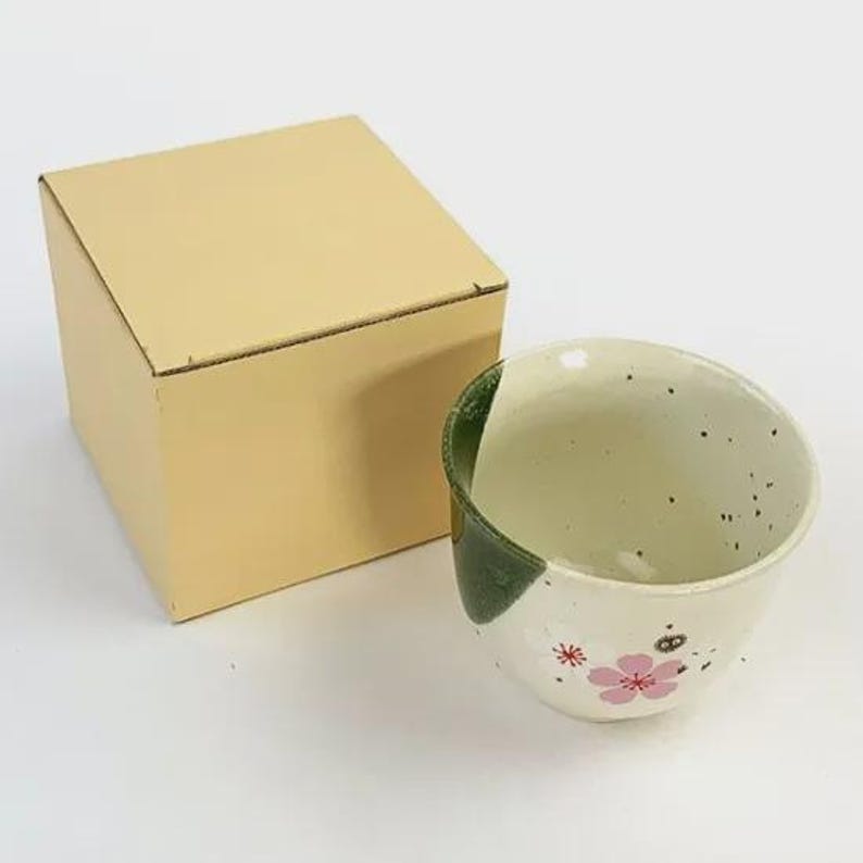 May include: A ceramic teacup with a white and green design, featuring pink floral accents and black speckles. The teacup is next to a beige cardboard box. The teacup has a unique, asymmetrical shape.