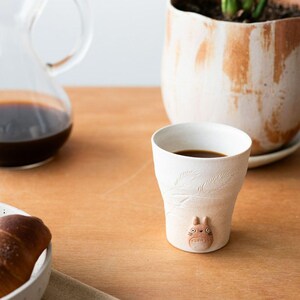 May include: A white ceramic coffee cup with a Totoro figure and leaf design. The cup is filled with dark coffee and sits on a wooden table. A glass carafe with coffee and a plant in a pot are in the background.