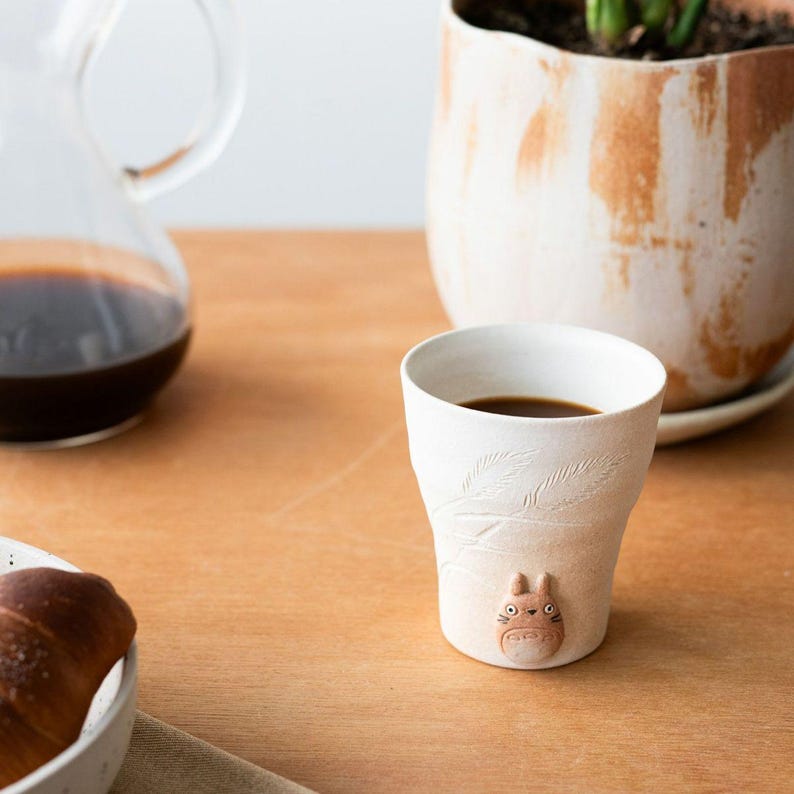 May include: A white ceramic coffee cup with a Totoro figure and leaf design. The cup is filled with dark coffee and sits on a wooden table. A glass carafe with coffee and a plant in a pot are in the background.