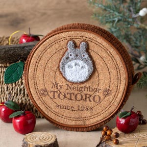 May include: Round cork coaster featuring a felt applique of Totoro from My Neighbor Totoro. The coaster has a brown corduroy edge and the text "My Neighbor TOTORO since 1988". Decorative apples and greenery surround the coaster.