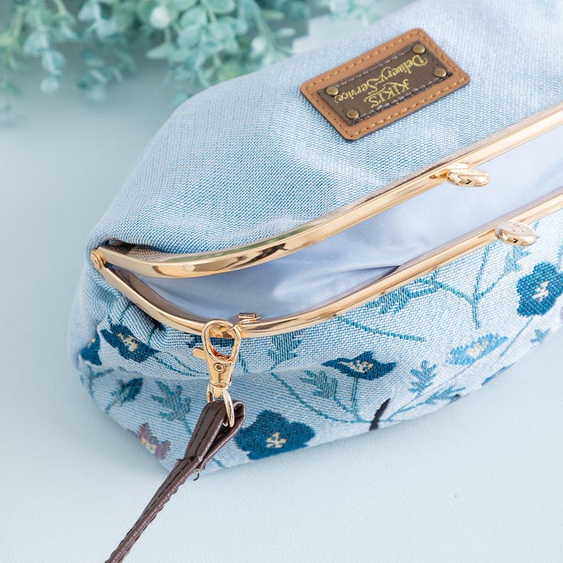 May include: A light blue denim clutch with a gold-colored clasp and a brown leather strap. The clutch features a floral pattern in shades of blue and a brown leather tag with the text "Kiki's Delivery Service".