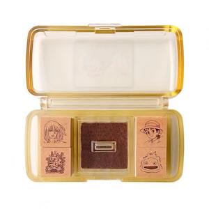 May include: A clear plastic stamp set with a gold-colored frame, containing four stamps with cartoon character illustrations and a brown ink pad. The stamps feature detailed drawings of anime characters.