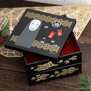 May include: Black square bento box with a red interior and a lid featuring a No-Face character, soot sprites, and Japanese text. The box has gold wave and geometric designs.