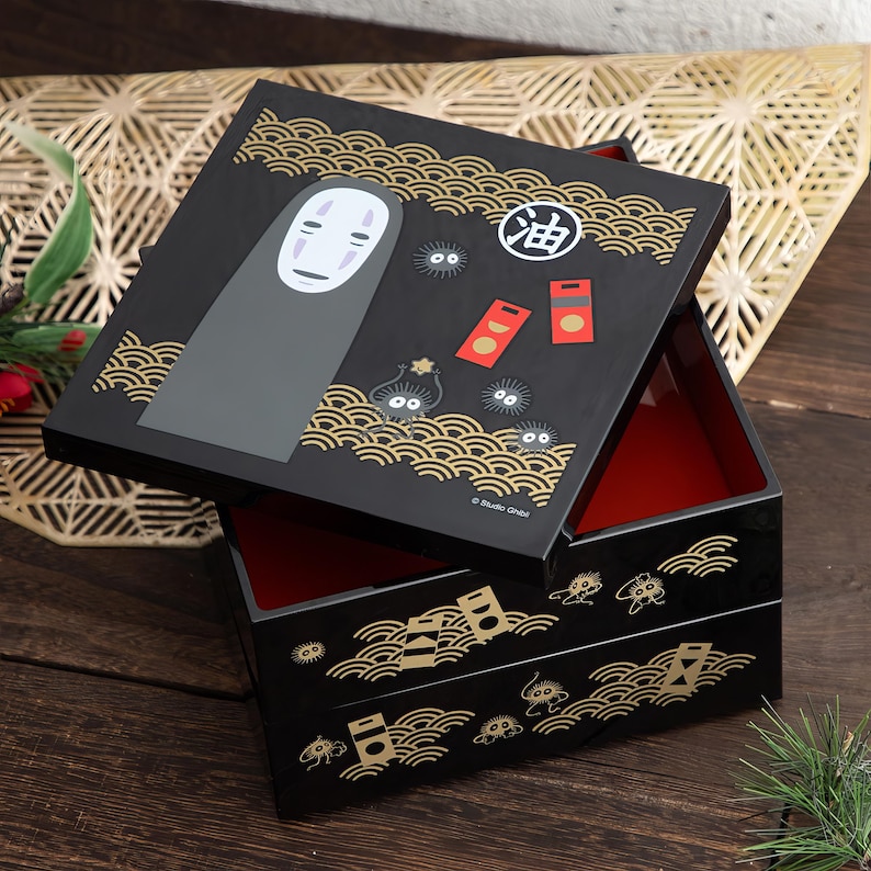 May include: Black square bento box with a red interior and a lid featuring a No-Face character, soot sprites, and Japanese text. The box has gold wave and geometric designs.