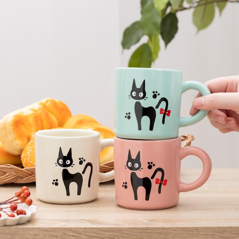 May include: Three ceramic mugs in light blue, cream, and pink, each featuring a black cat illustration with a red bow on the tail and small paw prints. The mugs have a rounded handle and a smooth finish, displaying a consistent cat graphic design.