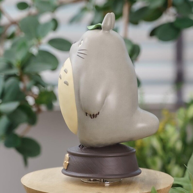 May include: A gray figurine of a Totoro character with a light yellow belly and a small green leaf on its head. The figurine has black whiskers and is mounted on a brown base. The background features green foliage.