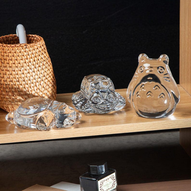 May include: Three clear glass figurines: a turtle, a turtle-shaped box, and a Totoro character. The figurines are displayed on a wooden shelf next to a woven basket. The turtle figurine is on the left, the turtle-shaped box is in the middle, and the Totoro is on the right.