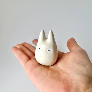 May include: A small, off-white ceramic figurine of a cartoon character with large, round eyes and two pointed ears. The figurine is held in a person's hand against a plain white background. The character has a simple, smiling face.