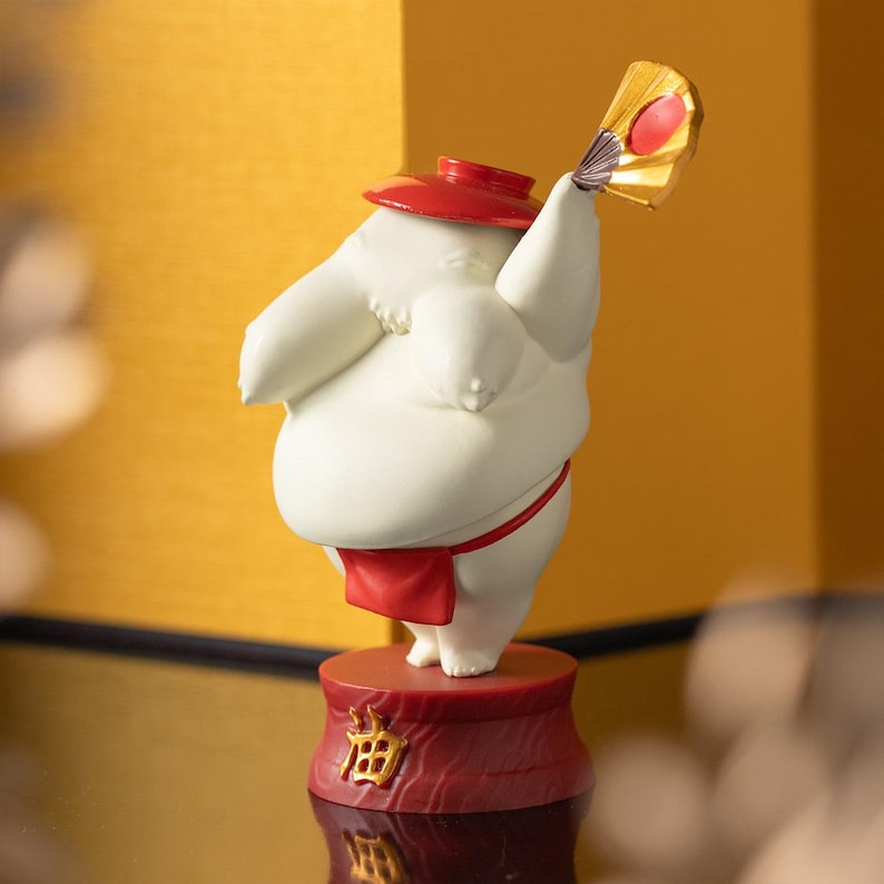 Peut inclure: Figurine blanche repr&eacute;sentant un lutteur de sumo avec un chapeau et un pagne rouges, tenant un &eacute;ventail dor&eacute; avec un accent rouge. La figurine repose sur une base rouge avec des caract&egrave;res japonais dor&eacute;s. Le fond est dor&eacute;.