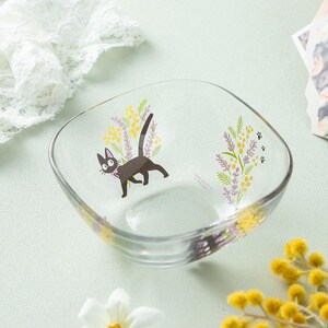 May include: A clear glass bowl featuring a printed graphic of a black cat with a pink collar, surrounded by yellow mimosa flowers and purple lavender sprigs. Small black paw prints are visible near the floral design on the curved glass surface.