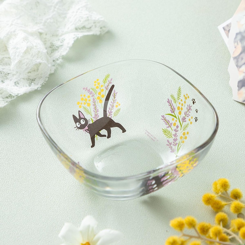 May include: A clear glass bowl featuring a printed graphic of a black cat with a pink collar, surrounded by yellow mimosa flowers and purple lavender sprigs. Small black paw prints are visible near the floral design on the curved glass surface.