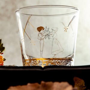 May include: A clear glass tumbler with a gold illustration of two figures and starbursts. The figures are holding a glowing object. The bottom of the glass has a gold filigree design. The glass is on a dark surface.