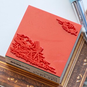 May include: A square red rubber stamp featuring a raised relief design of a windmill and clouds. The stamp is mounted on a wooden block with text printed on the side, including the words Made in China and Studio Ghibli Enterprises Inc.