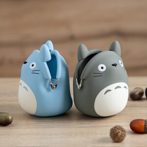 May include: Two coin purses shaped like cartoon characters, one light blue and one gray. Each has a white belly, black eyes, and a silver zipper. The purses are sitting on a wooden surface.