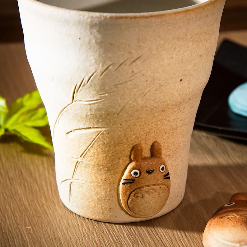 May include: Tan ceramic mug with a textured surface and a raised Totoro character design. The mug features etched leaf details and a rounded base. The mug is a light brown color and is suitable for hot or cold beverages.