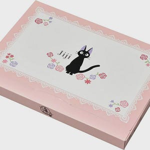 May include: A pink box with a white top featuring a black cat illustration with the word "Jiji". The top has a white lace border and floral accents in pink, purple, and blue. The box is closed.
