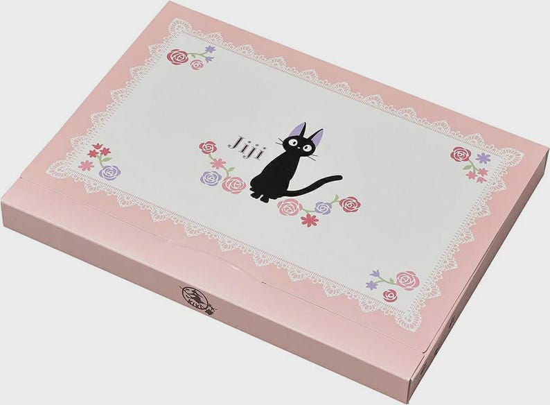 May include: A pink box with a white top featuring a black cat illustration with the word "Jiji". The top has a white lace border and floral accents in pink, purple, and blue. The box is closed.
