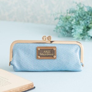 May include: A light blue canvas clutch purse with a gold metal frame and clasp. The purse has a brown leather patch with the text "Kikis Delivery Service" in gold lettering.