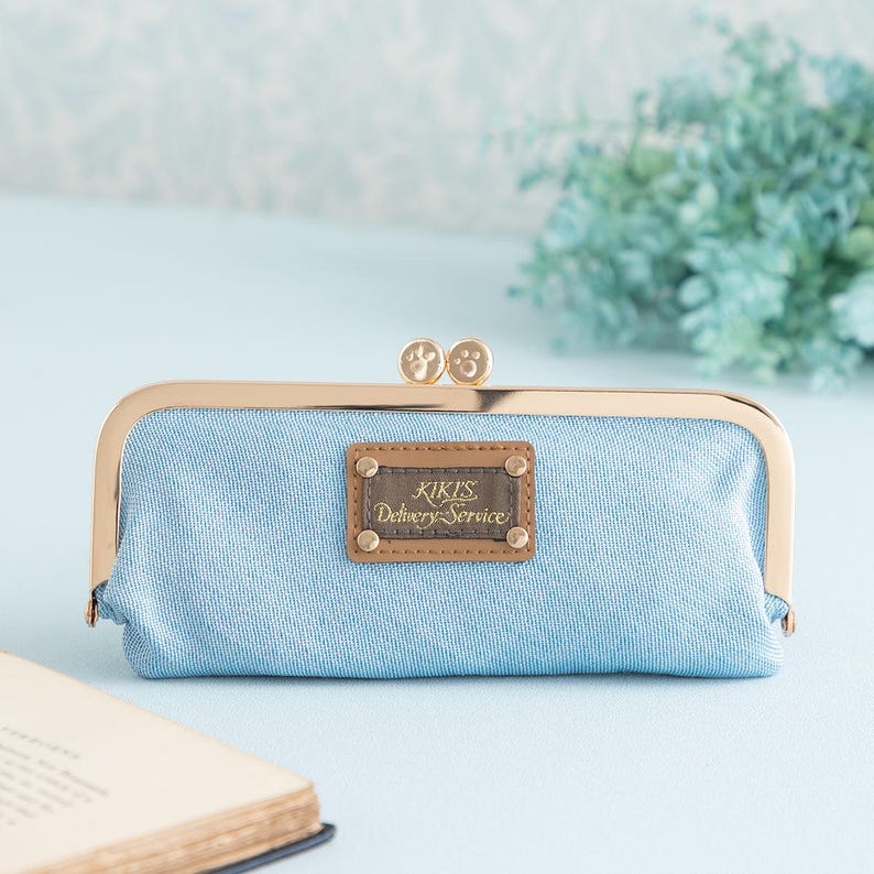 May include: A light blue canvas clutch purse with a gold metal frame and clasp. The purse has a brown leather patch with the text "Kikis Delivery Service" in gold lettering.