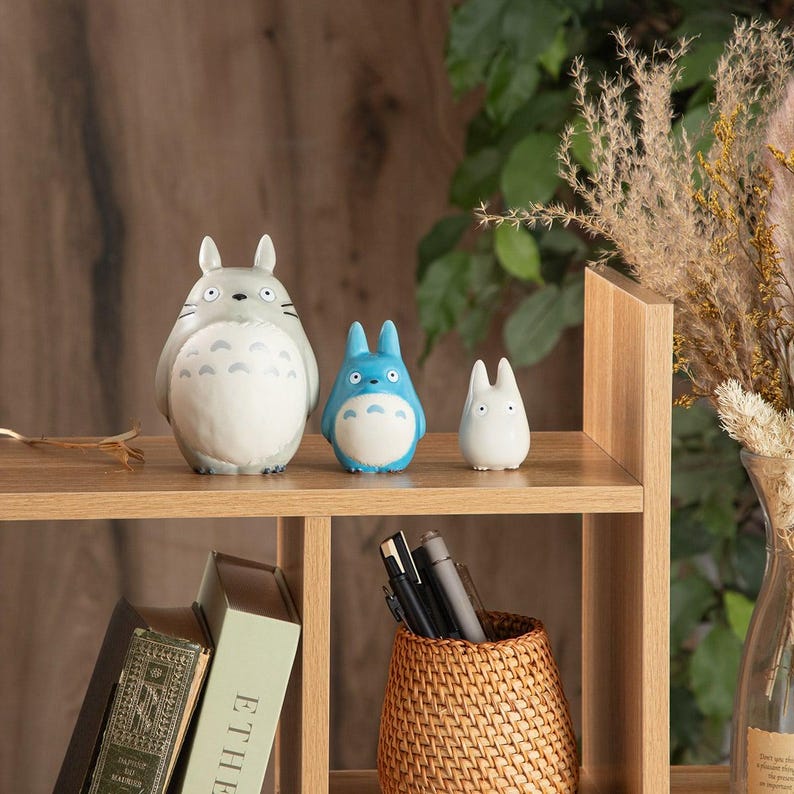 May include: Three figurines of the animated character Totoro in gray, blue, and white. The figurines are displayed on a wooden shelf with books and a woven basket. The background includes dried plants and a wooden structure.