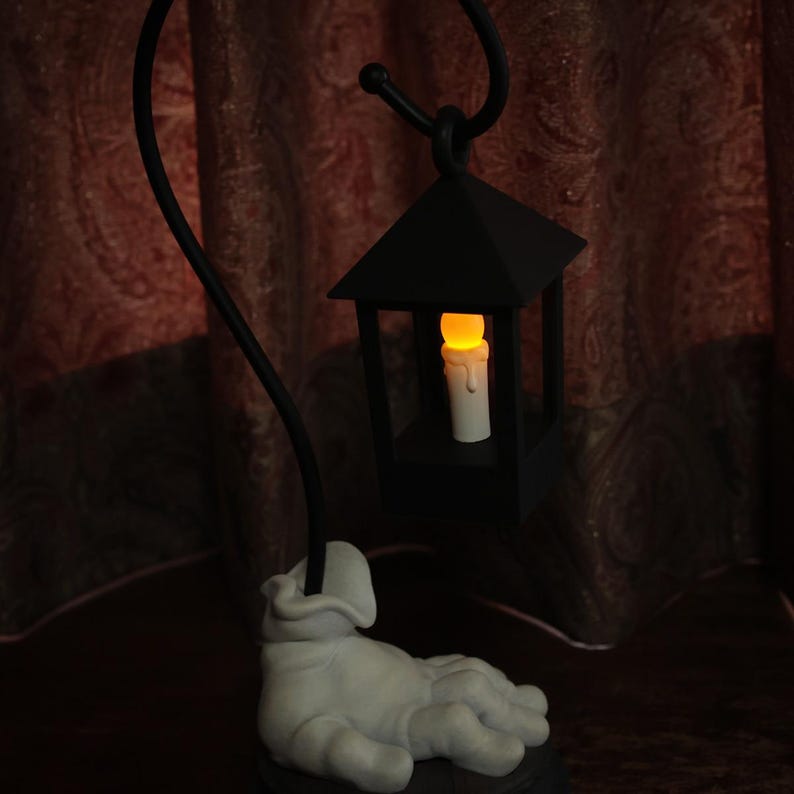 May include: A black metal lantern with a lit candle inside, suspended from a curved black stand. The base of the stand features a white, hand-shaped sculpture. The candle emits a warm, yellow glow.