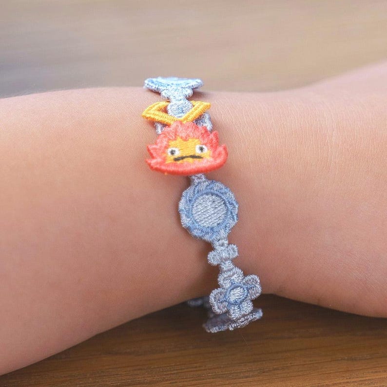 Op de afbeelding: Geborduurde armband met een stripfiguur met oranje en gele vlammen. De armband heeft een lichtblauwe denim-achtige uitstraling met een cirkelvormig en bloemig ontwerp. De armband wordt om de pols gedragen.