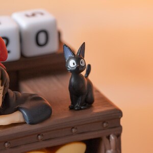 Pode incluir: Uma pequena figura de gato preto com olhos grandes e redondos e orelhas pontudas. O gato est&aacute; em uma superf&iacute;cie de madeira. O fundo inclui um bloco branco com o n&uacute;mero "0".