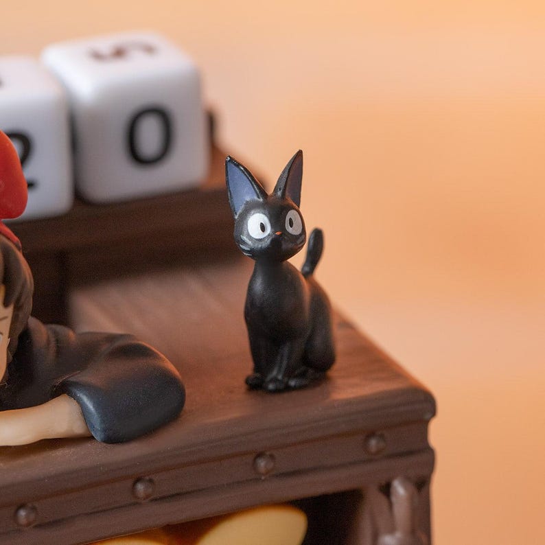 Pode incluir: Uma pequena figura de gato preto com olhos grandes e redondos e orelhas pontudas. O gato est&aacute; em uma superf&iacute;cie de madeira. O fundo inclui um bloco branco com o n&uacute;mero "0".