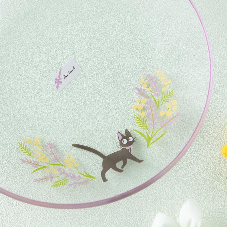 Op de afbeelding: Een helder glazen bord met een illustratie van een zwarte kat met een roze halsband, omringd door gele en paarse bloemtakjes. Een klein wit rechthoekig label met de tekst Kiki's Delivery Service is op het oppervlak bevestigd.