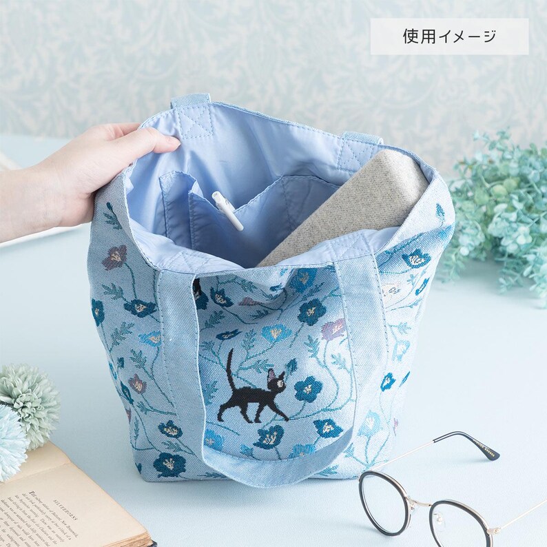 May include: A light blue tote bag with a black cat and floral pattern. The bag has a zippered pocket on the inside and is made of a soft, durable fabric.