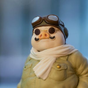 May include: A charming figurine of a pig in an aviator outfit. The pig is wearing a brown leather helmet and goggles, a light green jacket, a white scarf, and small round glasses. The pig has a pink snout and black moustache.