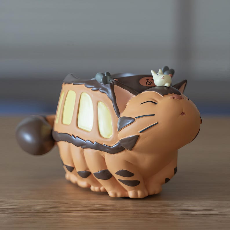 May include: A tan and brown decorative planter shaped like a cat-bus from the animated film, with a small white figure on top. The planter has a round tail and a series of rectangular windows. The cat-bus has black stripes and details.