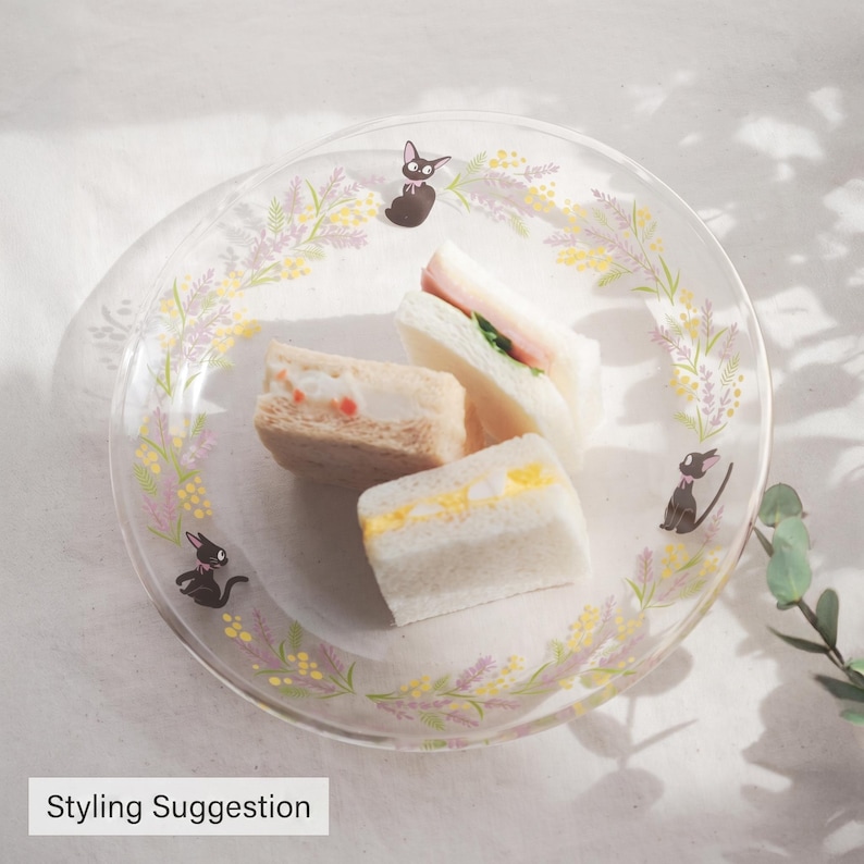 May include: A clear glass plate decorated with a floral wreath pattern of yellow and purple flowers and three black cat illustrations, holding three small square sandwiches with visible fillings of egg, ham, and a creamy spread.
