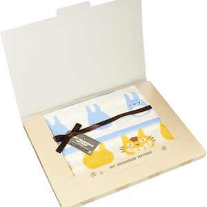 May include: A white towel with blue and yellow cartoon characters, presented in a gift box. The towel features a blue border and a brown ribbon. The box has the text "MY NEIGHBOR TOTORO".