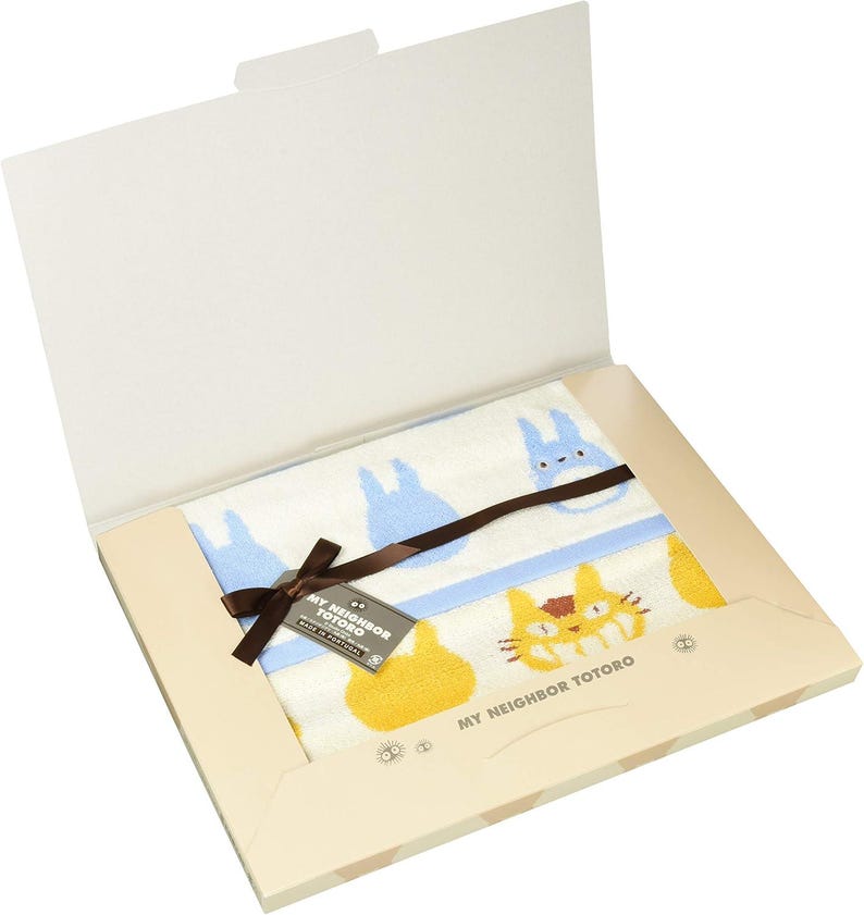 May include: A white towel with blue and yellow cartoon characters, presented in a gift box. The towel features a blue border and a brown ribbon. The box has the text "MY NEIGHBOR TOTORO".