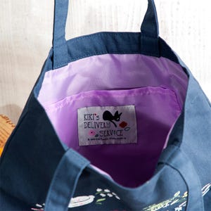 K&ouml;nnte beinhalten: Eine dunkelblaue Tragetasche mit lilafarbener Innenausstattung und einer kleinen Tasche. Die Tasche hat ein wei&szlig;es Etikett mit schwarzem Text, auf dem "Kiki's Delivery Service" steht, und eine schwarze Katzensilhouette. Die Tasche hat einen Blumendruck mit einem wei&szlig;en Teller und Blumen.